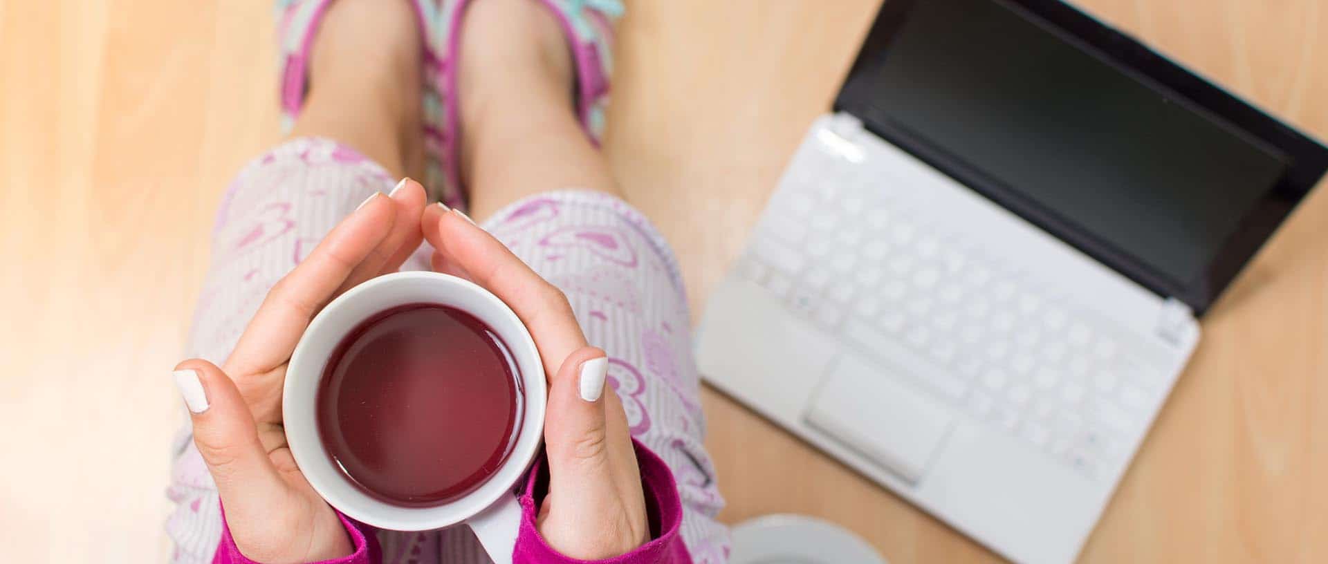 Freelance copywriter holding cup of tea, wearing pyjamas and slippers, with computer on the floor